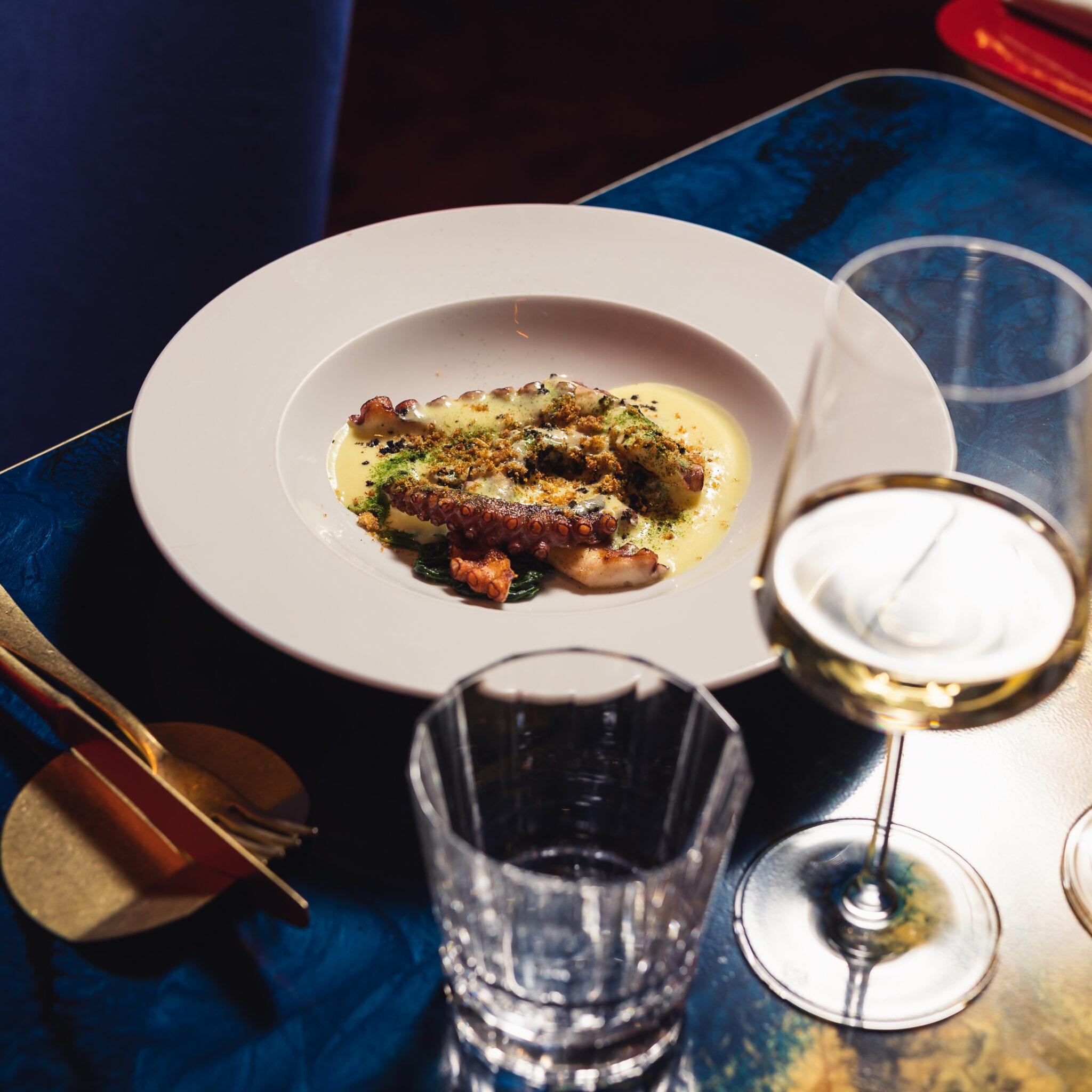 Un delizioso piatto di polpo a cena, accompagnato da un bicchiere di vino bianco e uno di acqua. Il polpo è servito con una salsa delicata e spezie.